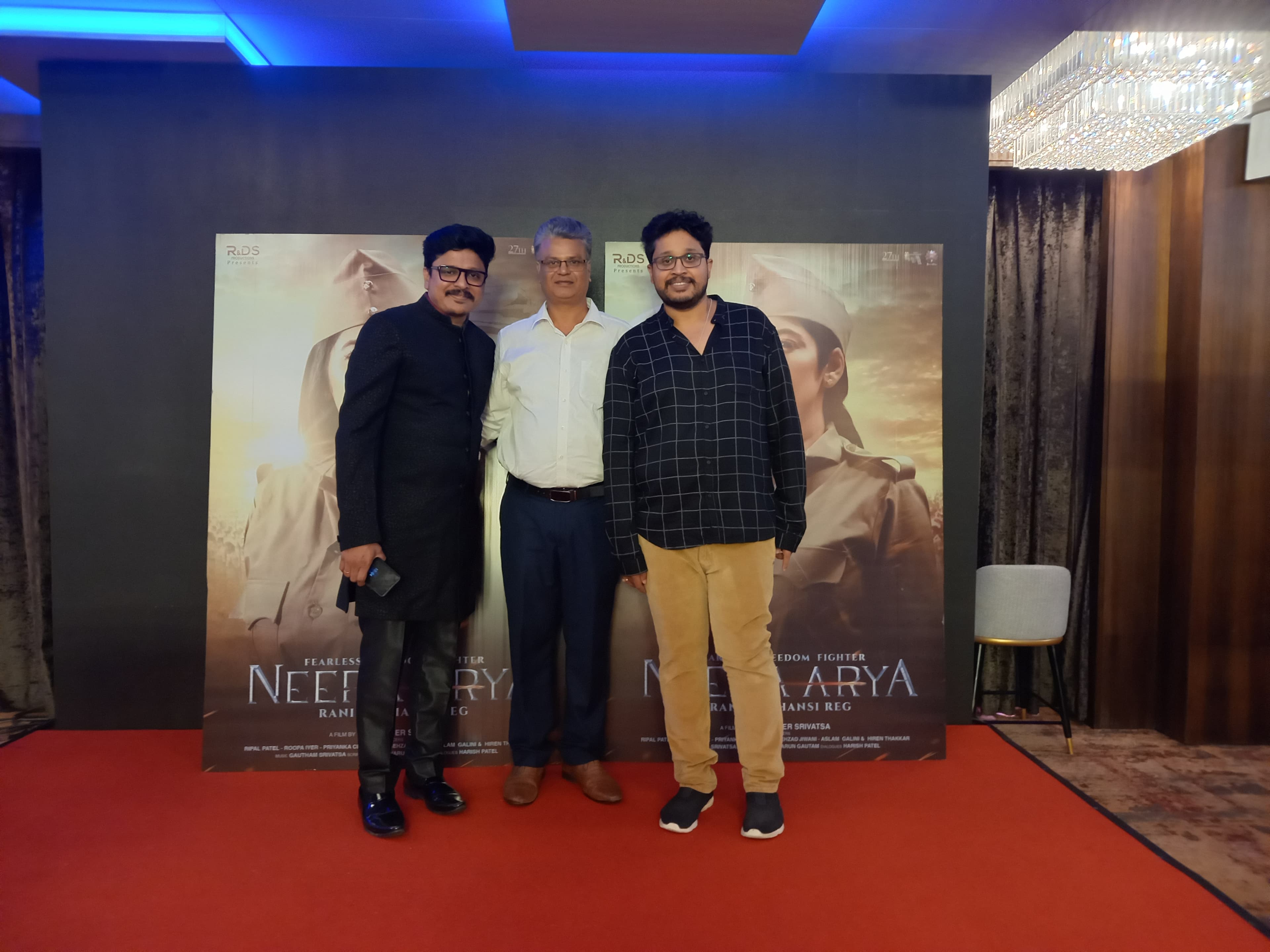 Three men on a red carpet in front of Neera Arya film posters